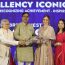 Four individuals standing together, with two men and two women. One man is handing a bouquet of flowers to a woman wearing a beige traditional outfit. The background displays the words "Excellency Iconic Awards" and "Recognizing Achievement at Bizz expo Jaipur - Inspiring Success."